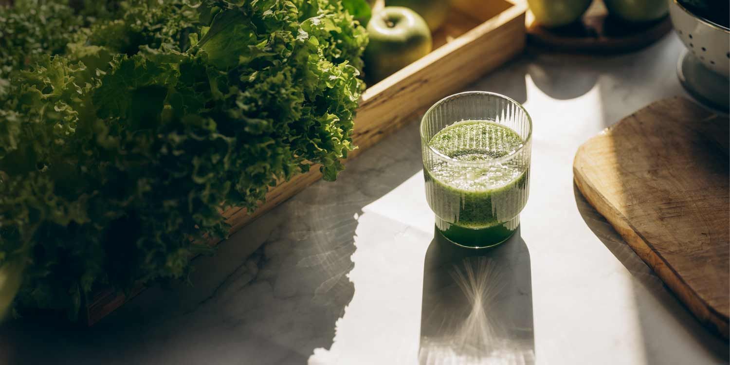 A glass of green smoothie lit by sunlight, next to kale and apples