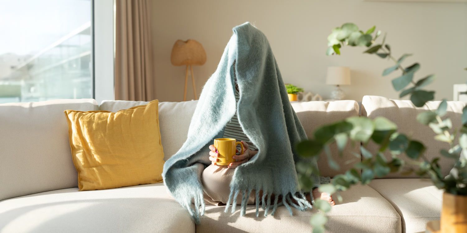 Person på soffa med filt över sig, håller en gul kopp i morgonljus
