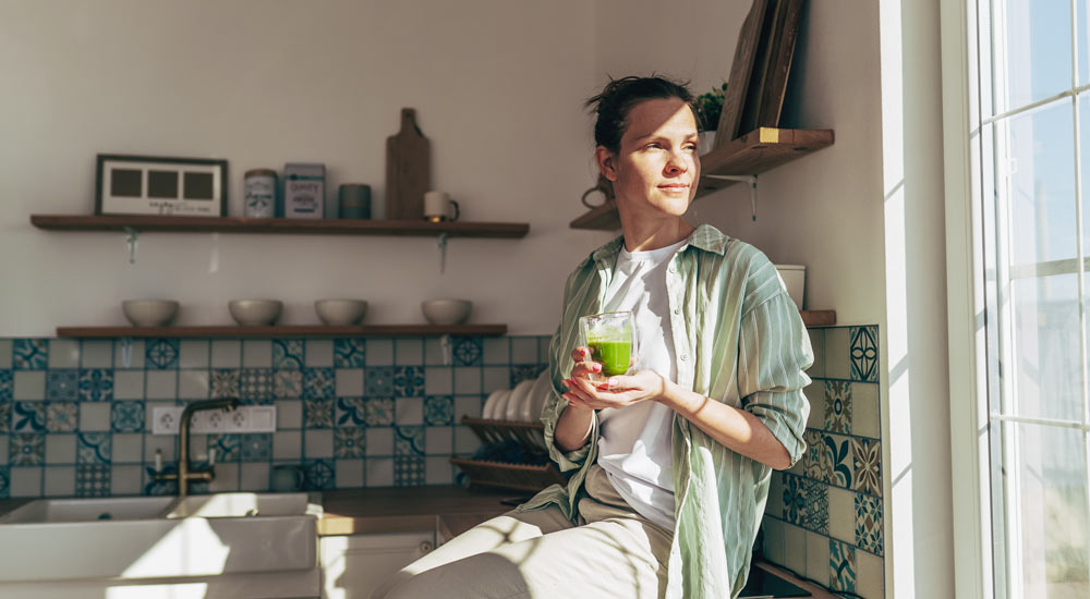Woman sitting on a kitchen counter in sunlight, drinking a green smoothie