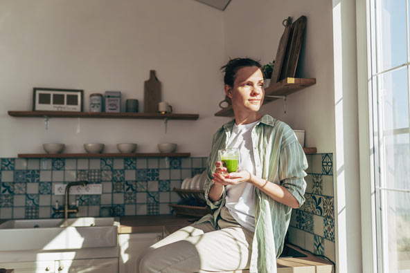 Woman sitting on a kitchen counter in sunlight, drinking a green smoothie