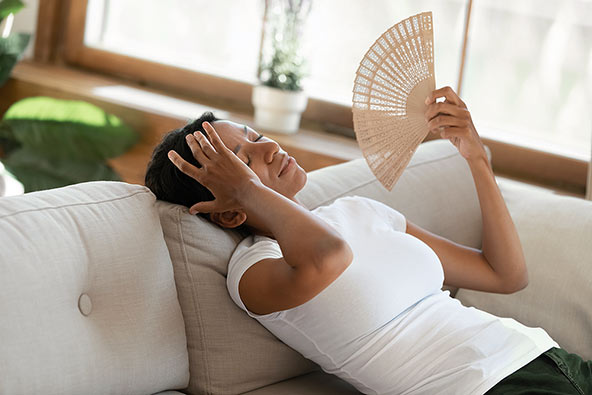 Trött och varm kvinna sitter i en soffa och fläktar sig med en solfjäder