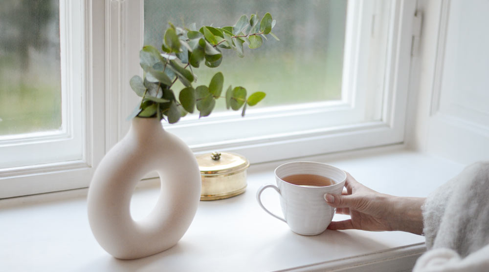 Kopp med varmt te i handen, placerad vid fönster i lugn och naturlig hemmiljö.