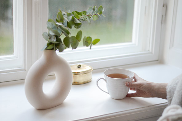 Kopp med varmt te i handen, placerad vid fönster i lugn och naturlig hemmiljö.