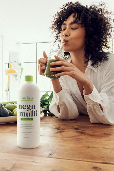Kvinna sitter och dricker en smoothie med en flaska Närokällan Mega Multi flytande multivitamin bredvid sig