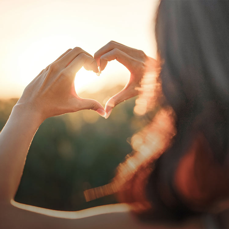 Woman holding up her hands in the shape of a heart agains the sun set