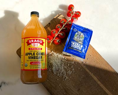 Äppelcidervinäger från Bragg, havssalt från Celtic Sea Salt och kvisttomater på en skärbräda med ljus bakgrund.