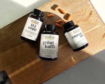 Tre kosttillskott från Närokällan på träbräda: D3 4000, grönt kaffe och Curcumin. Några orange kapslar är utspridda.