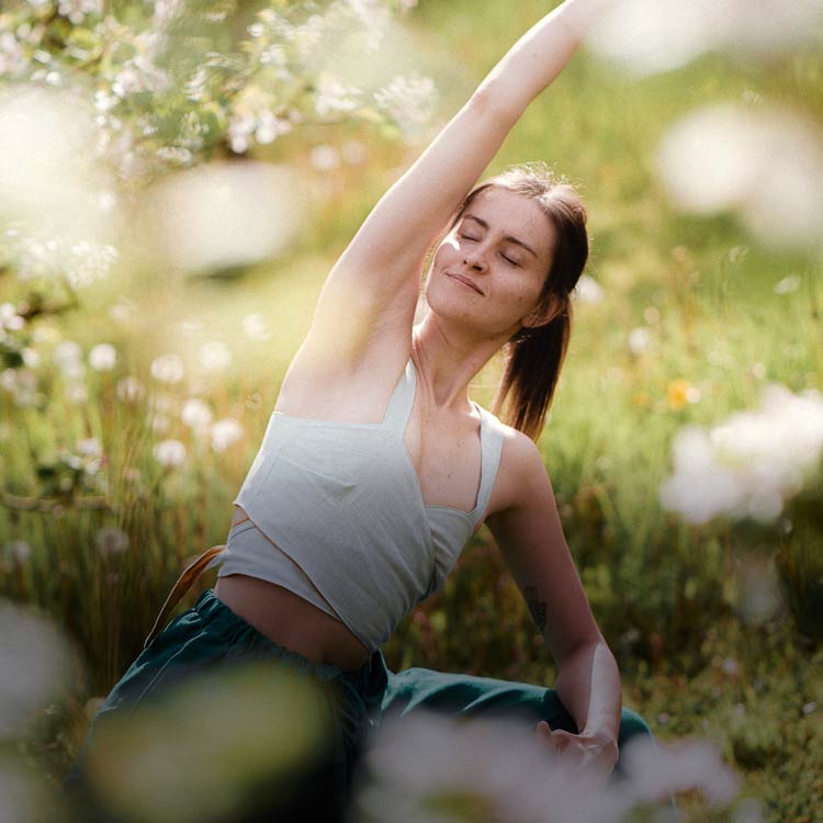 Kvinna i grönt linne sträcker sig i yogaposition på en sommaräng med ljusa blommor i bakgrunden.