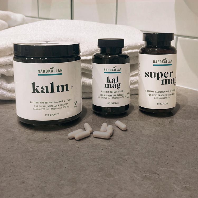 Tre burkar från Närokällan på badrumsbänk: Kalm+, KalMag och SuperMag, med vita kapslar framför och hopvikta handdukar i bakgrunden.